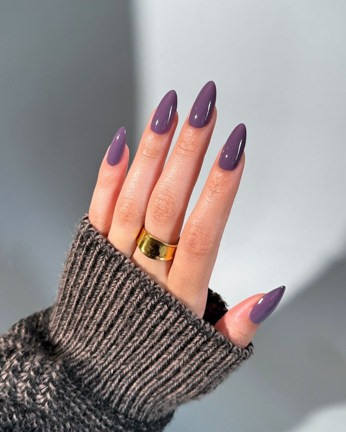 Unhas pintadas com esmalte roxo, mão com anel dourado e suéter cinza.