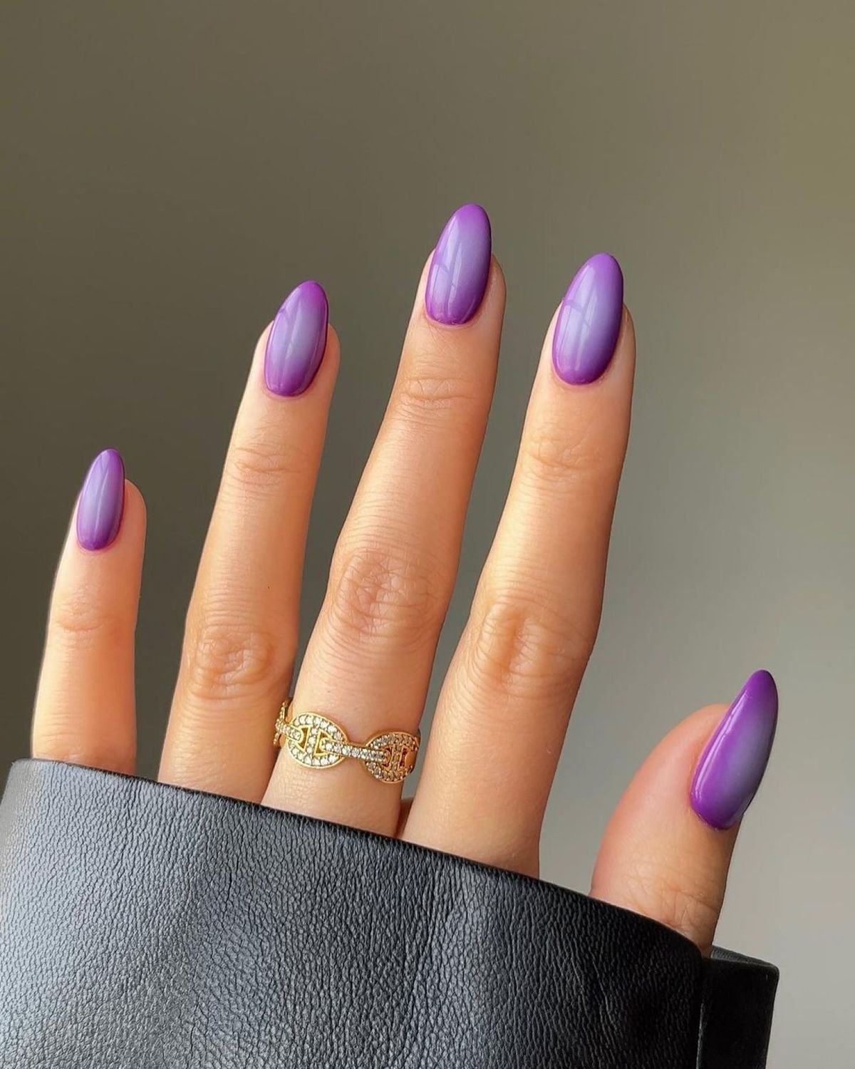 Unhas longas pintadas com esmalte roxo, destacadas por um anel dourado e pulseira preta.