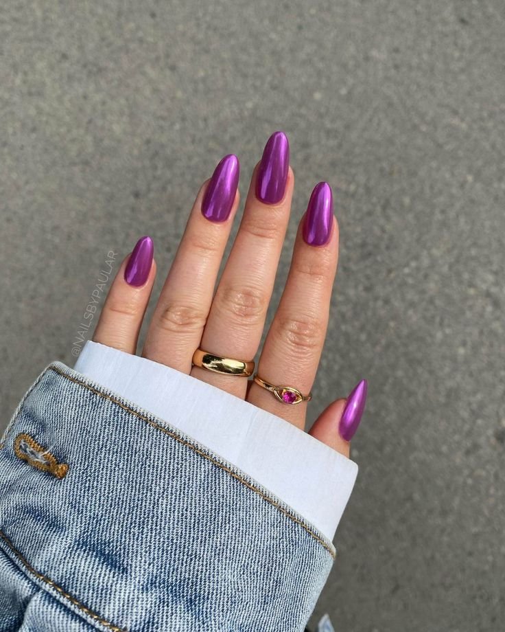 Unhas com esmalte roxo brilhante em mão com anéis dourados e manga de jaqueta jeans.