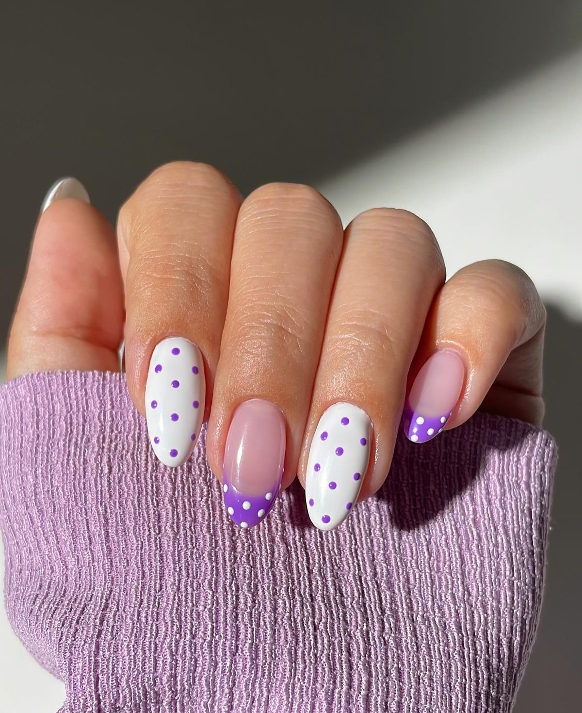 Unhas com esmalte roxo em bolinhas e francesinha sobre fundo branco e lilás.