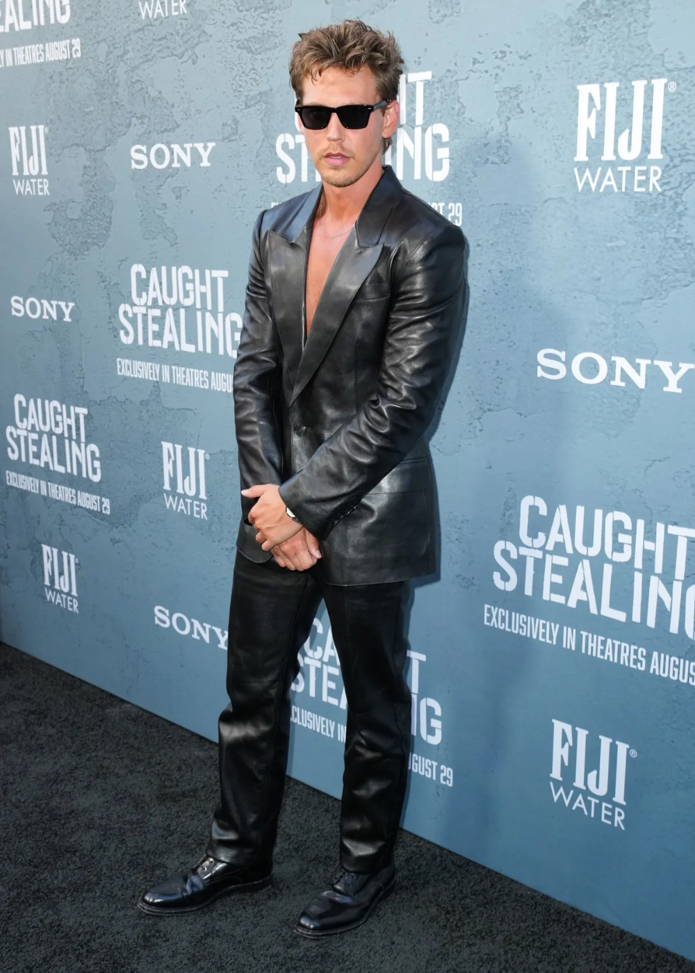 Austin Butler usa um terno de couro preto e óculos escuros em frente a um painel azul.