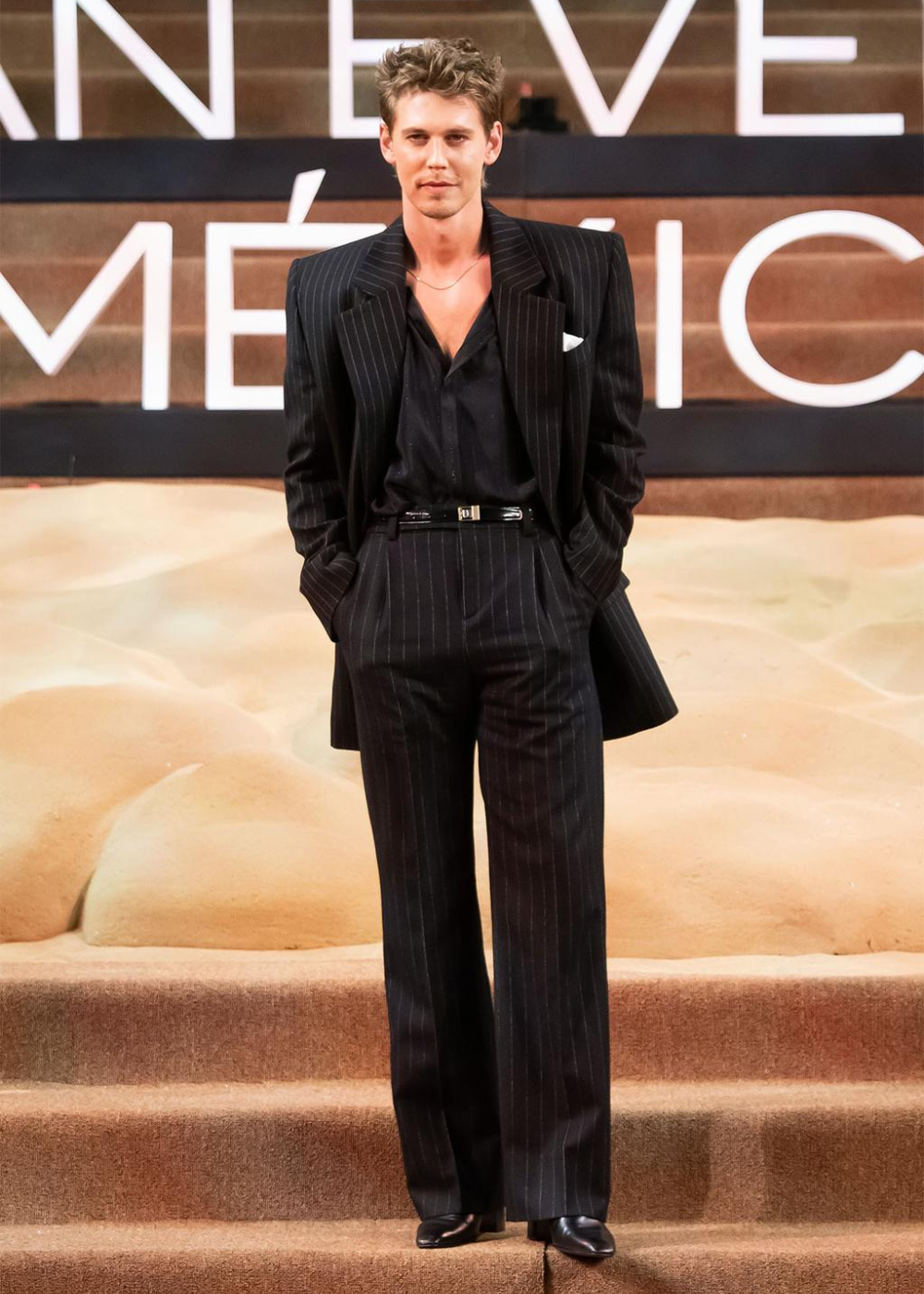 Austin Butler usando um terno risca de giz preto enquanto posa em pé com os braços para trás em frente a um painel.