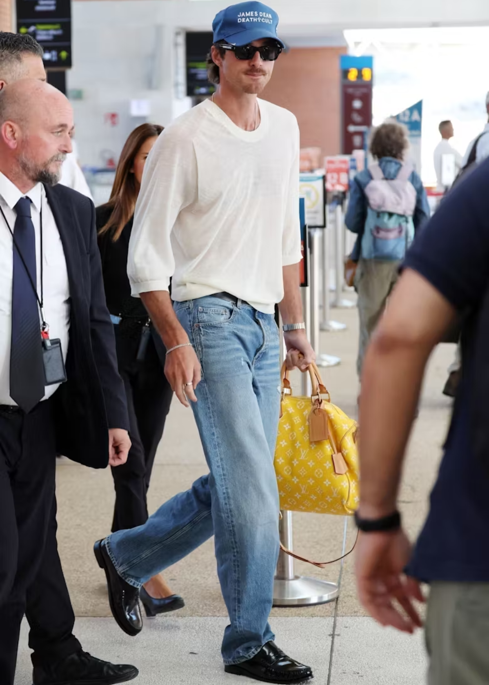 Jacob Elordi de jeans, blusa branca, boné azul e óculos escuros, carregando bolsa amarela em aeroporto.