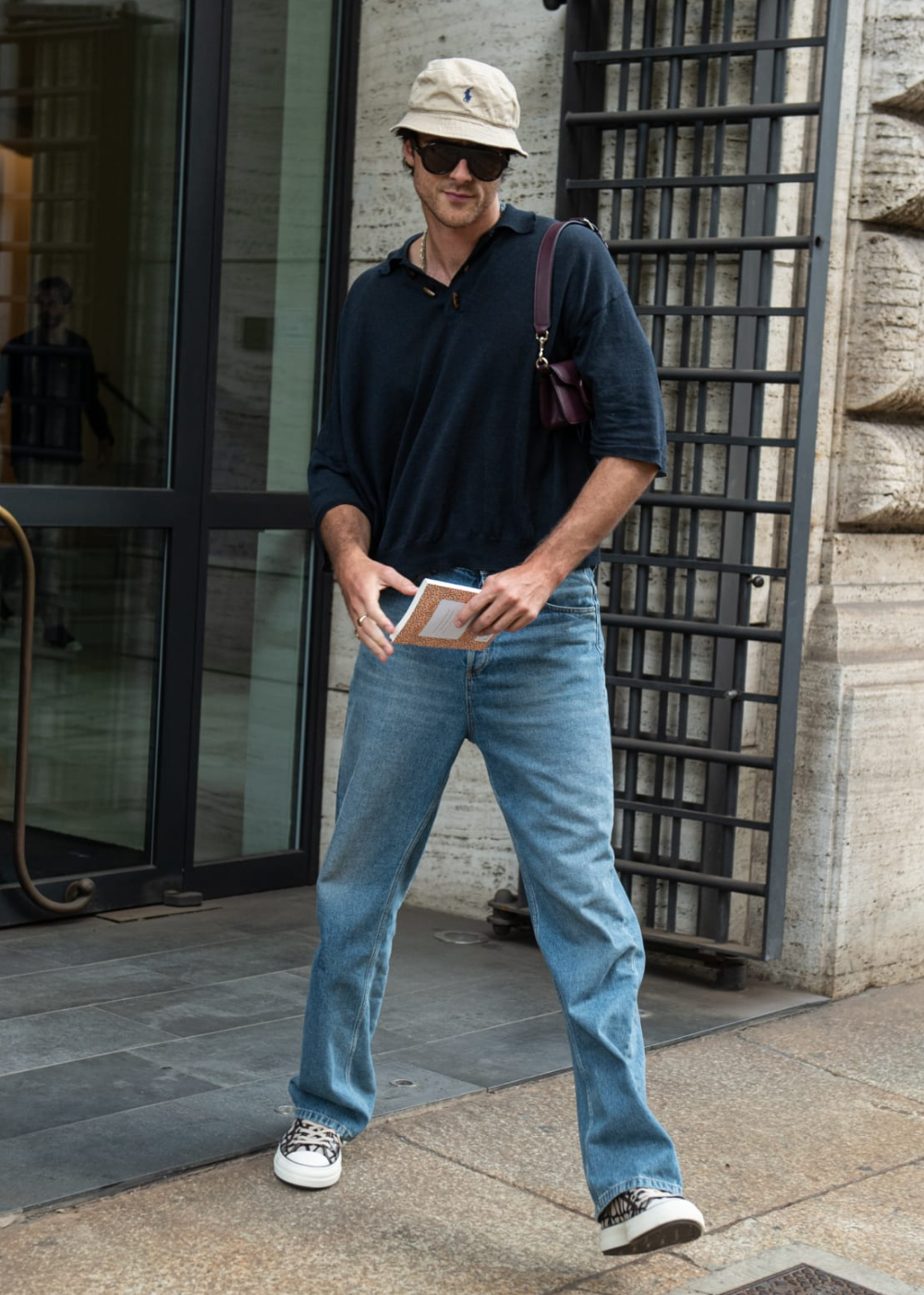 Jacob Elordi de boné bege, óculos escuros e camisa azul caminha pela rua segurando um livro.