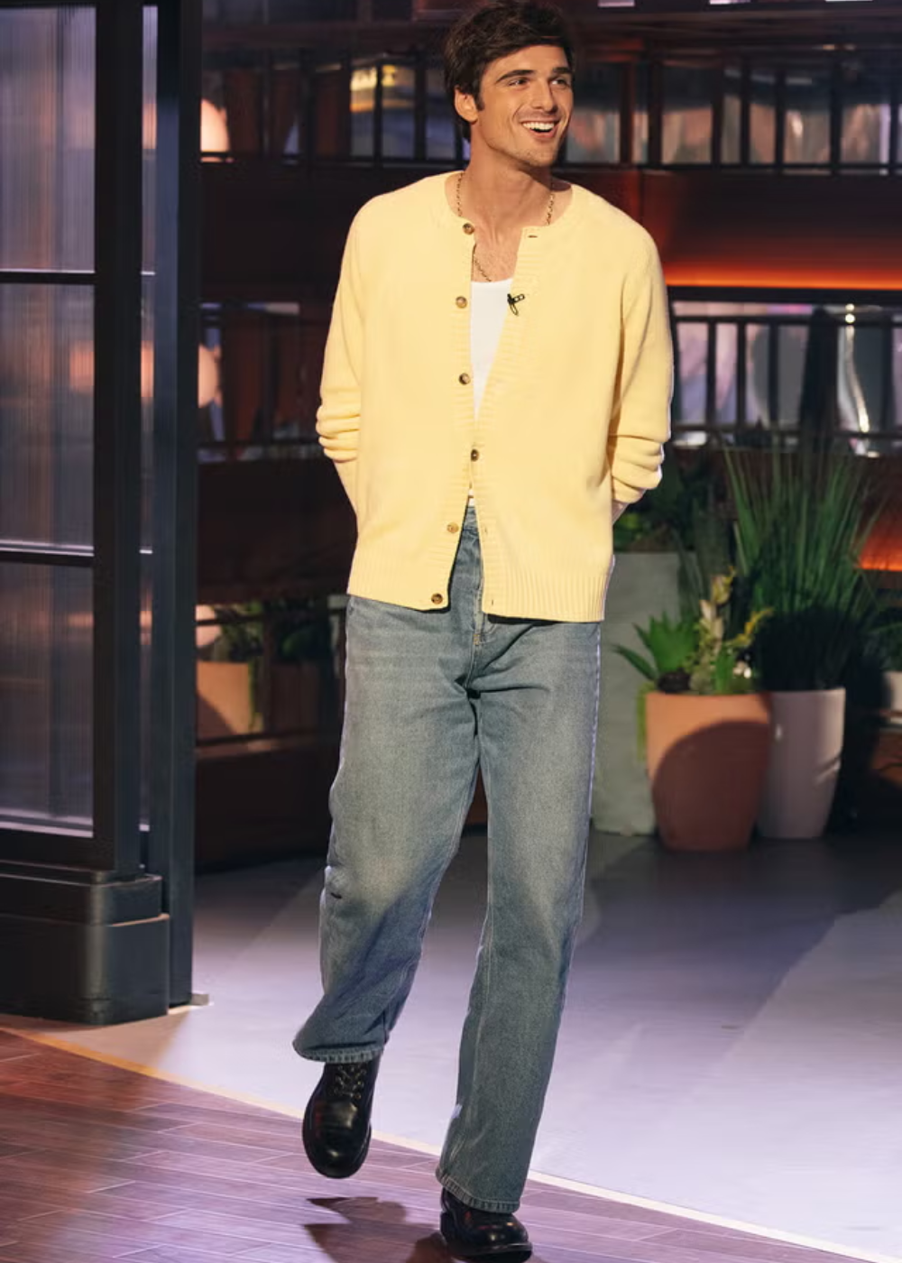 Um homem de suéter amarelo e jeans caminha em um estúdio iluminado com plantas ao fundo.