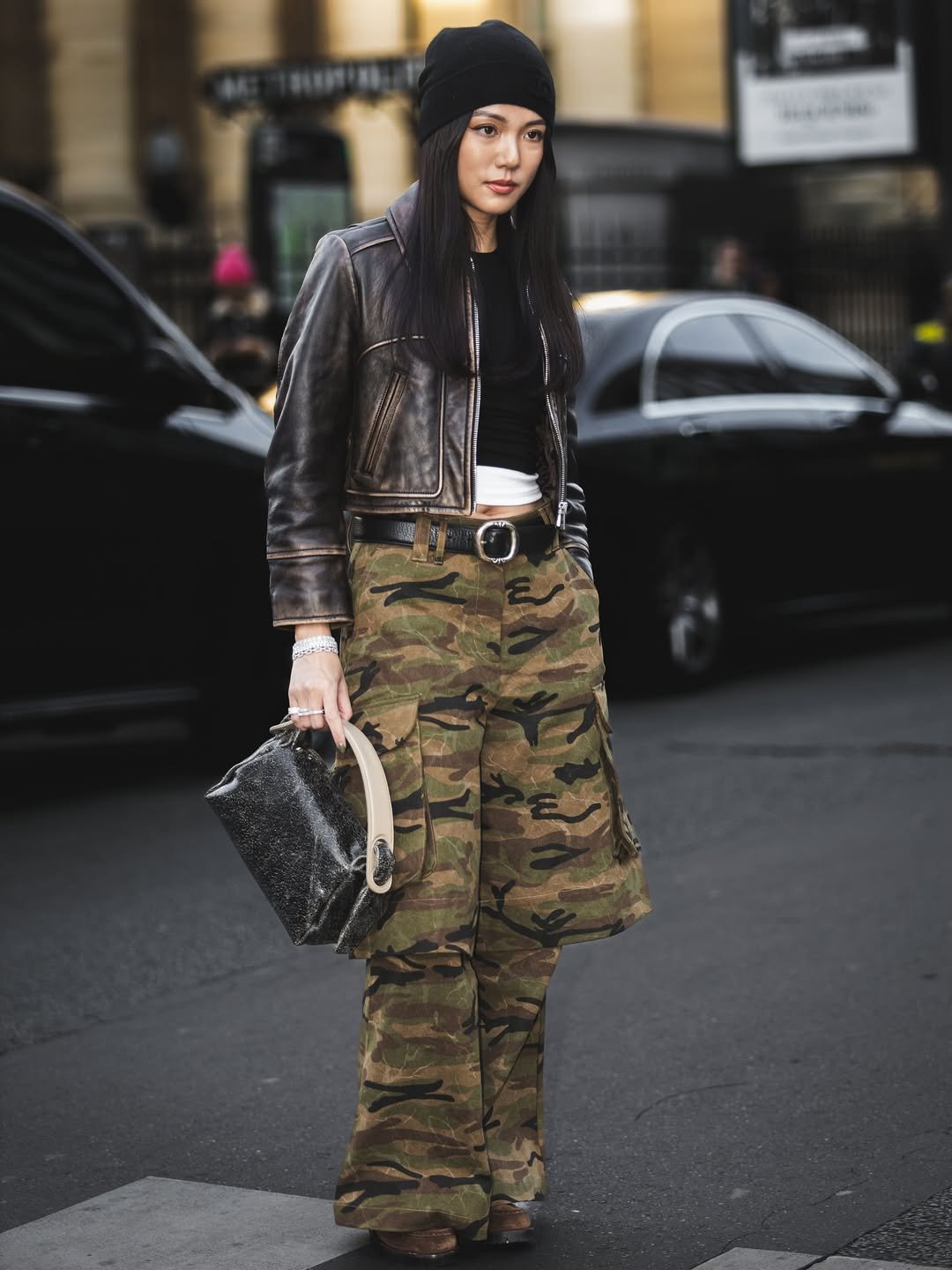Pessoa com calça de padrão camuflado, casaco de couro e bolsa, em uma rua urbana.