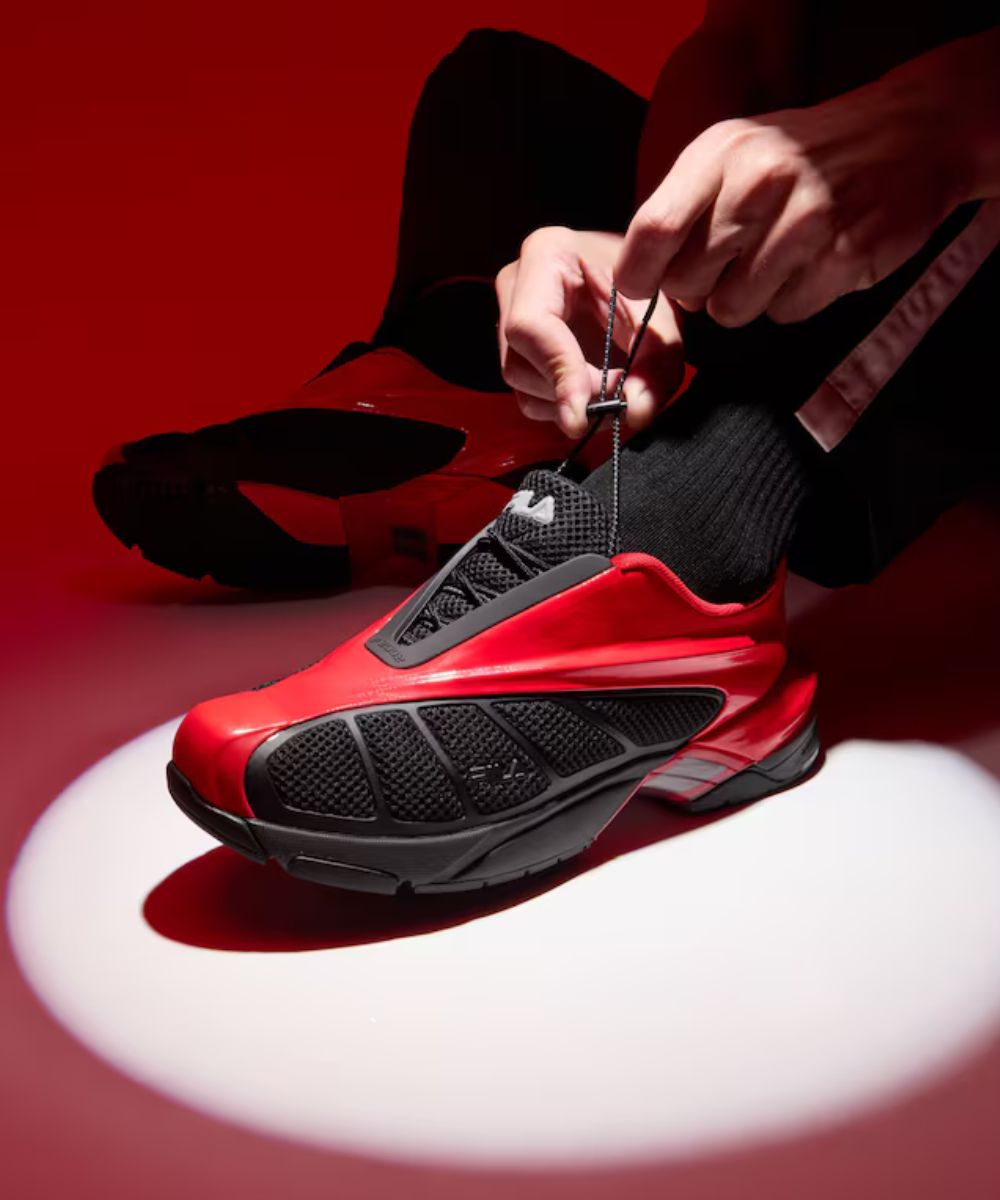 Replica 2002 de Fila. Tênis vermelho com detalhes pretos sendo amarrado, em fundo vermelho. Exibe design moderno.