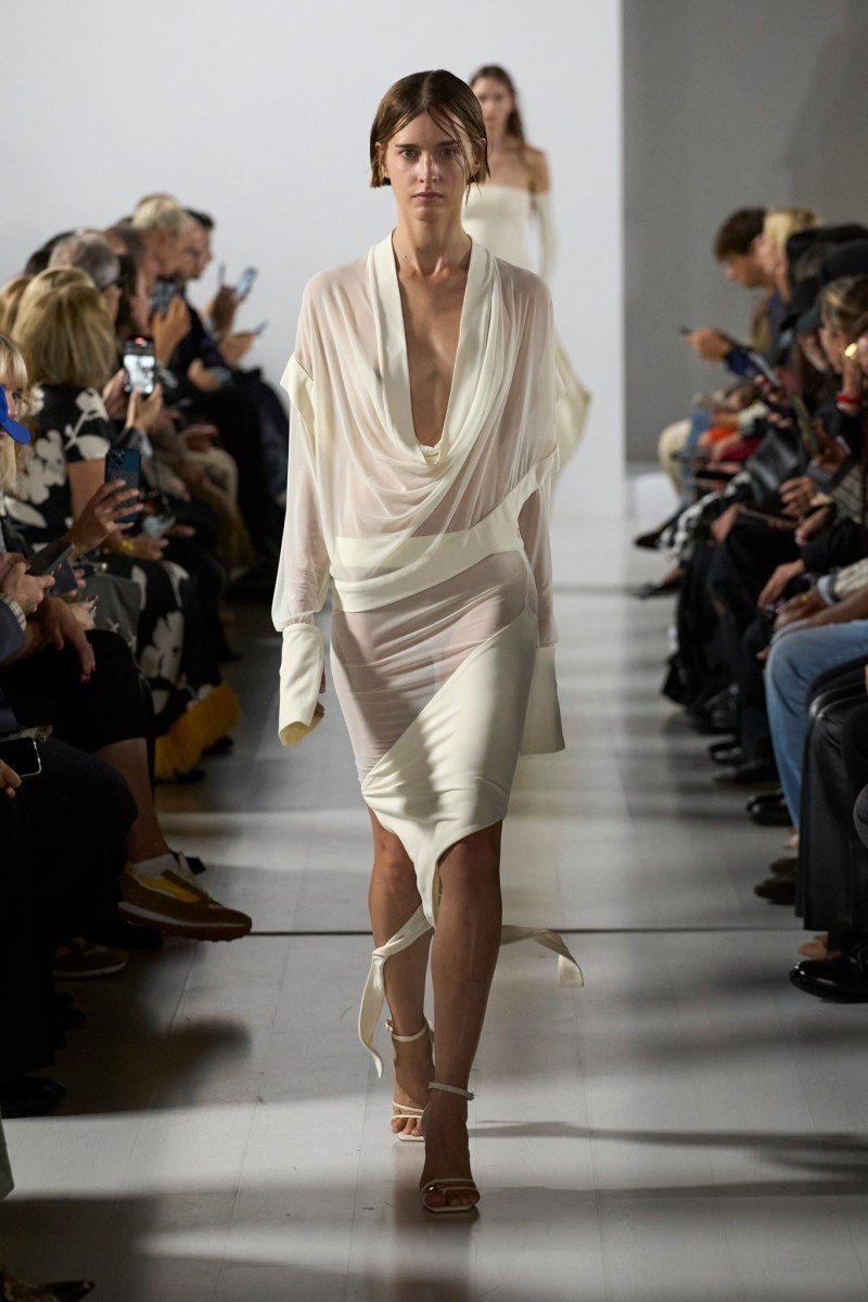 Desfile na Milan Fashion Week SS26 com modelo em vestido branco translúcido, cercado por espectadores.