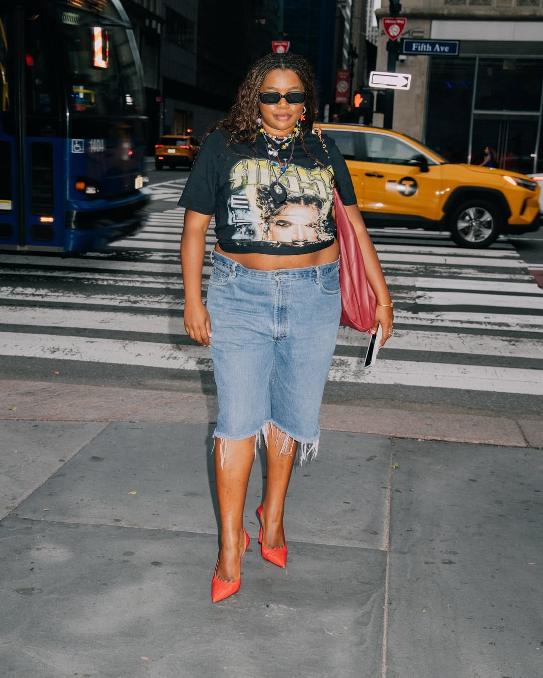 Pessoa com uma graphic tee em uma rua movimentada, usando shorts jeans e sapatos vermelhos, carregando uma bolsa vermelha.