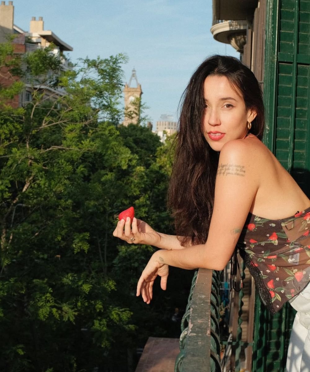 Mulher de cabelo ondulado segura um morango na varanda, com vista para árvores e uma torre ao fundo.