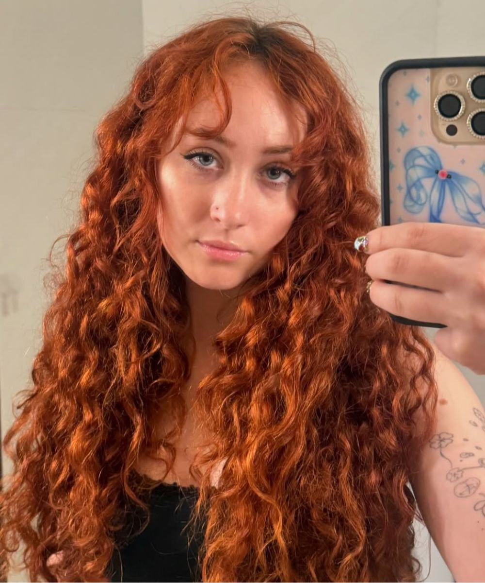 Pessoa com cabelo ondulado ruivo, tirando selfie no espelho. Há uma tatuagem visível no braço.