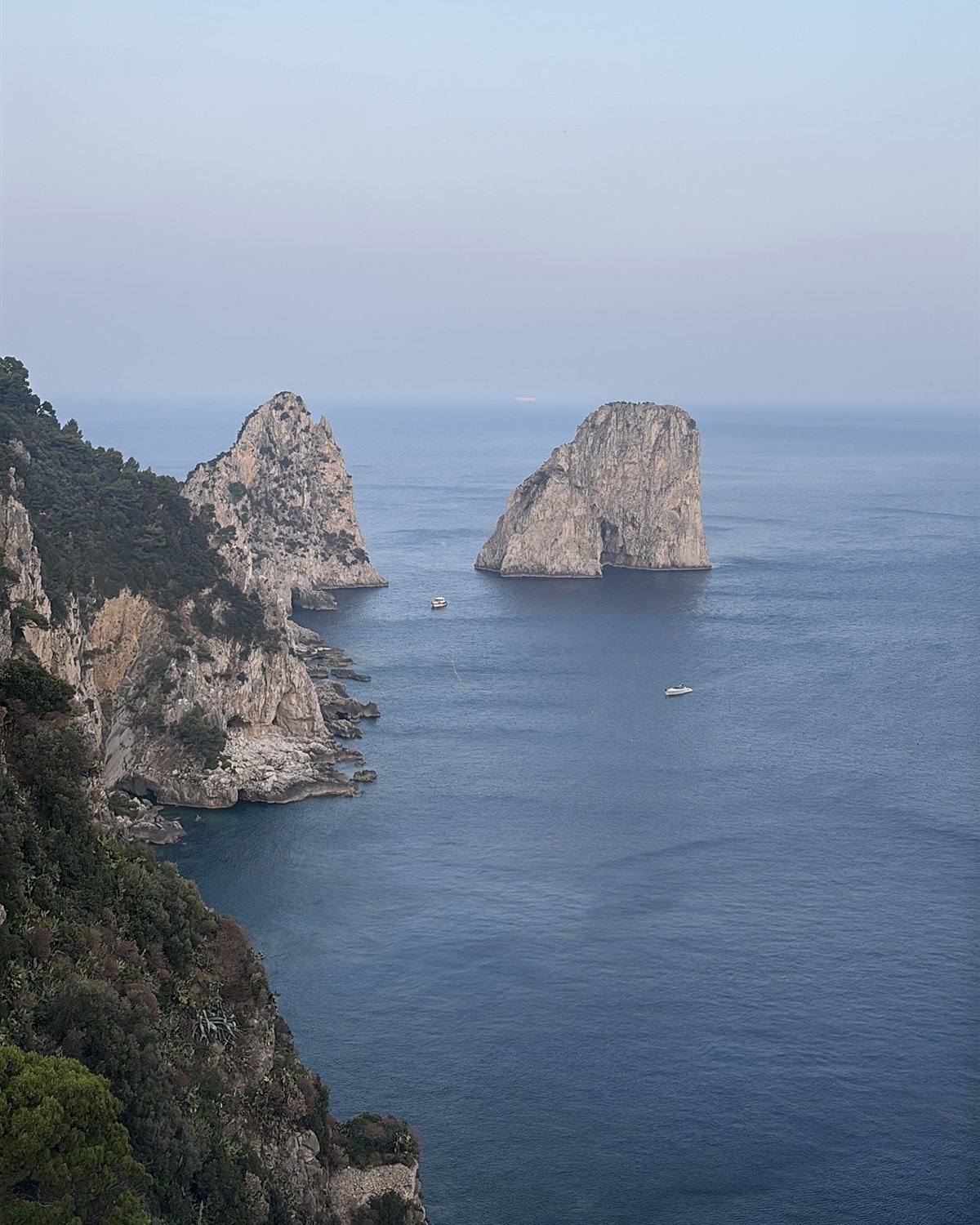 Mar azul claro com rochas imponentes emergindo da água e duas embarcações ao fundo. Vegetação cobre as falésias.