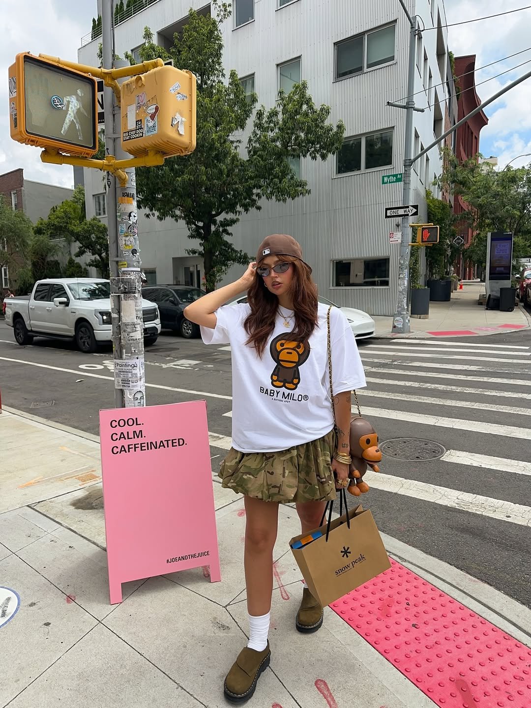 Pessoa com camiseta branca e saia camuflada em esquina urbana, segurando sacola e boneco.
