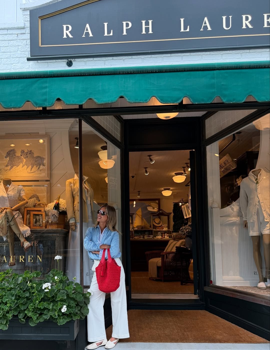 Mulher em frente a loja Ralph Lauren, com estilo preppy, segurando bolsa vermelha. Manequins e decoração interior visíveis.