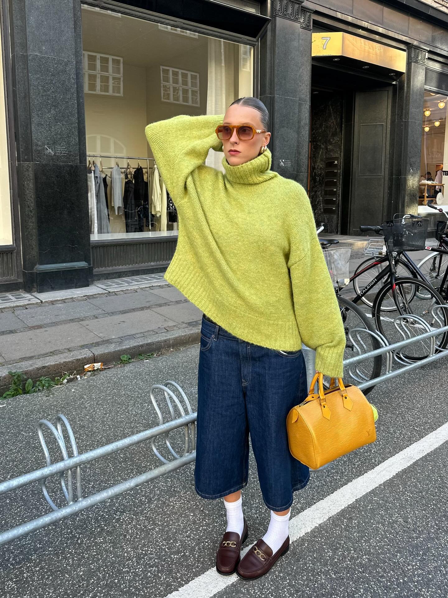 Pessoa com suéter amarelo, calça jeans larga e loafers marrons, segura bolsa amarela.