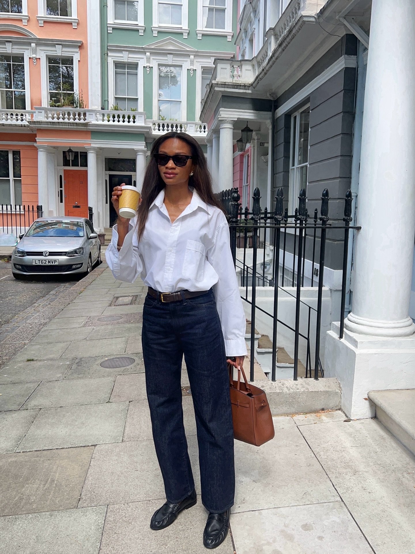 Mulher com jeans e loafer preto segura café. Ao fundo, prédios coloridos e bolsa marrom na mão.