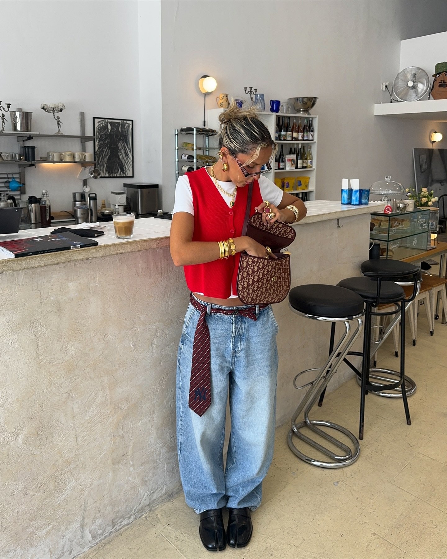 Pessoa com colete vermelho usa jeans folgado e loafer preto. Ela está olhando dentro de uma bolsa em um café moderno.