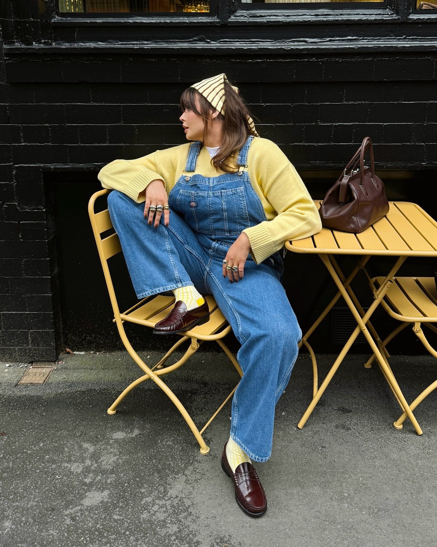 Pessoa em macacão jeans, suéter amarelo e loafers, sentada em cadeira amarela ao lado de mesa com bolsa marrom.