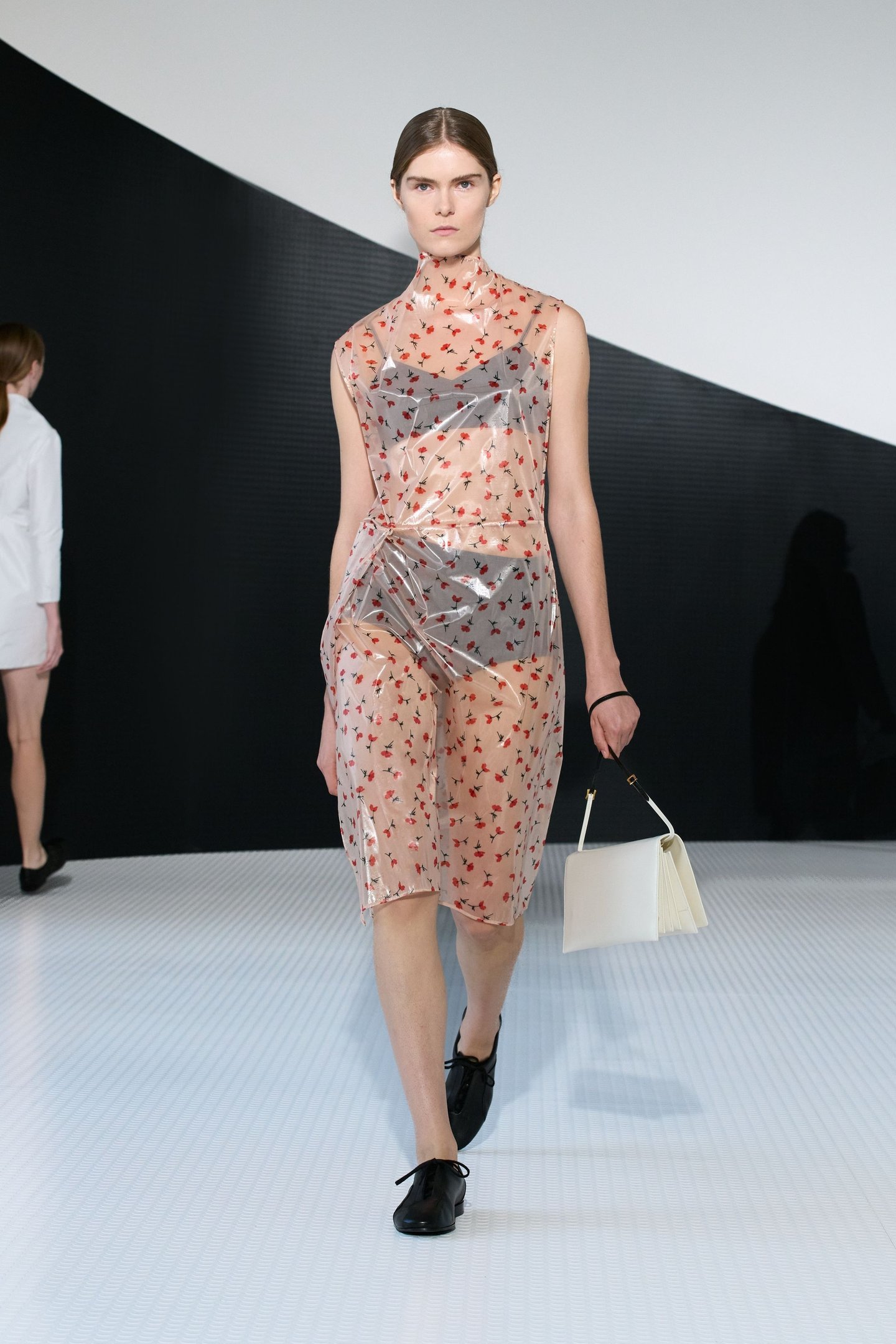 Modelo desfila com vestido transparente estampado e bolsa branca. Milan Fashion Week SS26.
