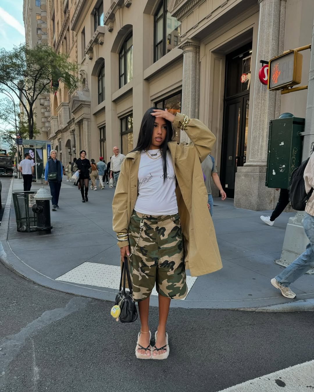 Jovem na calçada usando bermuda camuflada, casaco bege e segurando bolsa preta, com prédio ao fundo.
