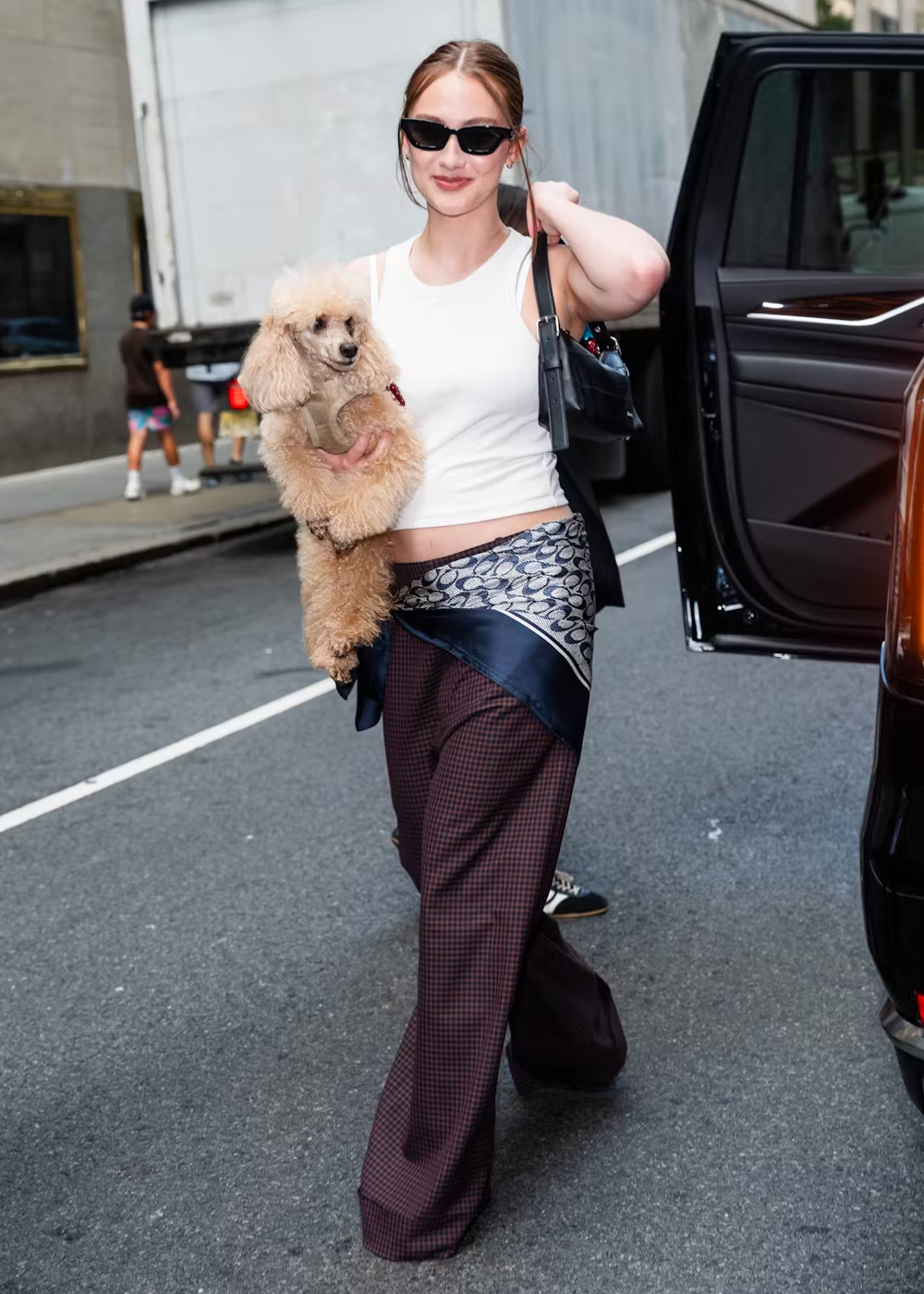 Lola Tung sorridente com óculos escuros carrega um poodle em uma rua movimentada, saindo de um carro preto.