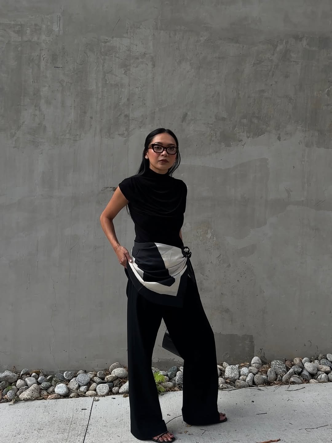 Pessoa posando com roupa preta e lenço amarrado na cintura, em frente a parede cinza.