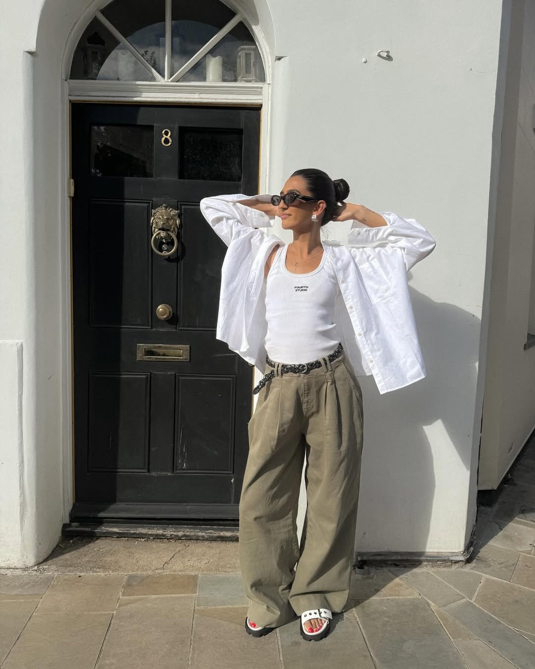 Pessoa de óculos escuros e look com camisa branca e top, em frente a uma porta preta.