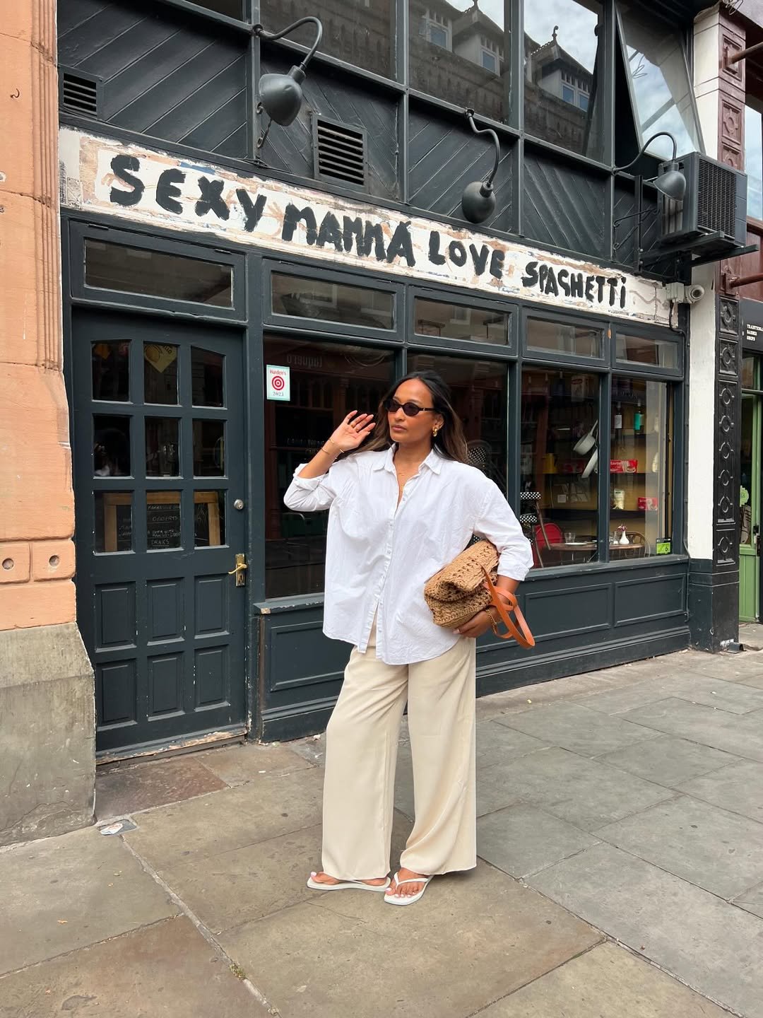 Mulher de óculos escuros veste look com camisa branca e calça bege, em frente a uma loja.