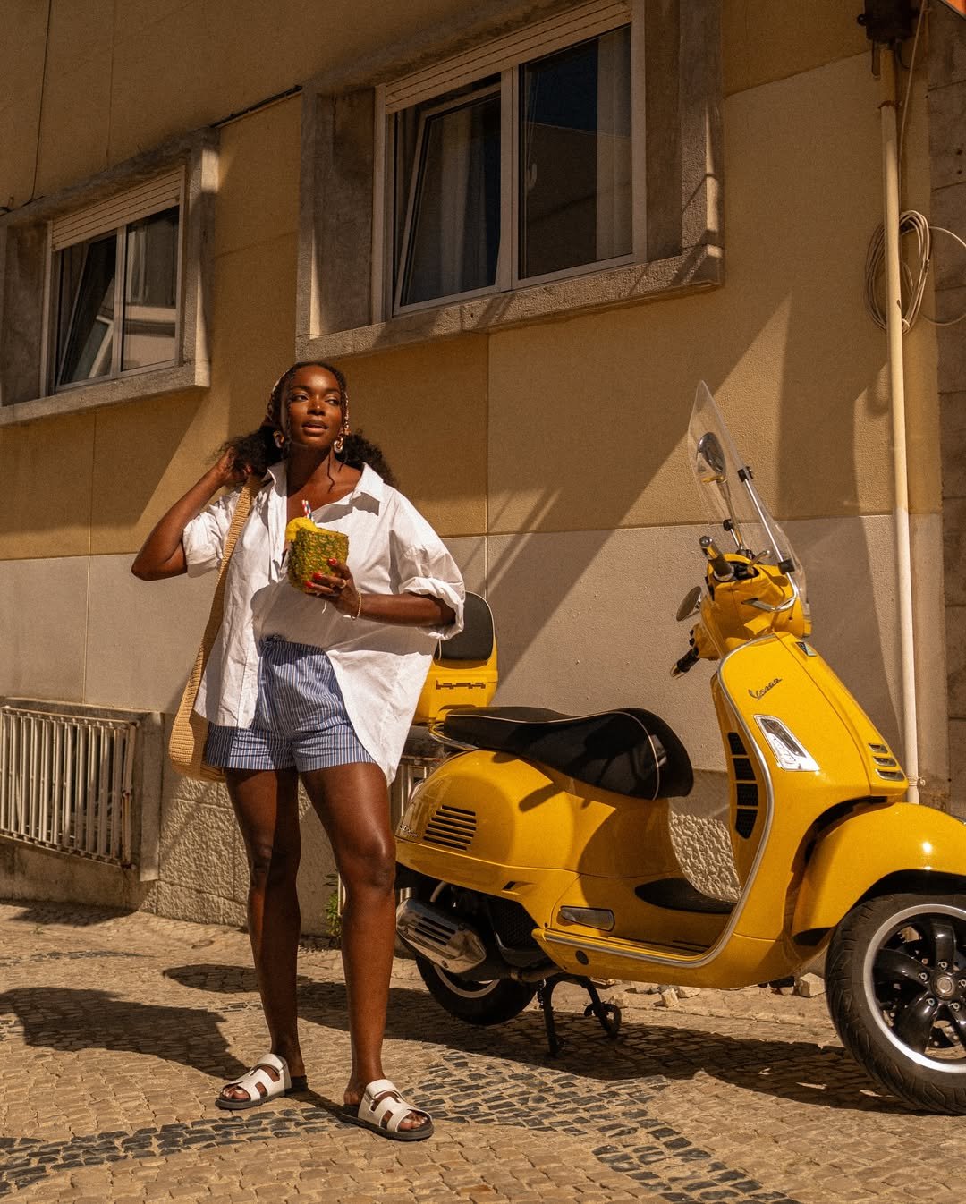 Mulher ao lado de uma scooter amarela, usando look com camisa branca. Segura bebida e bolsa de palha.