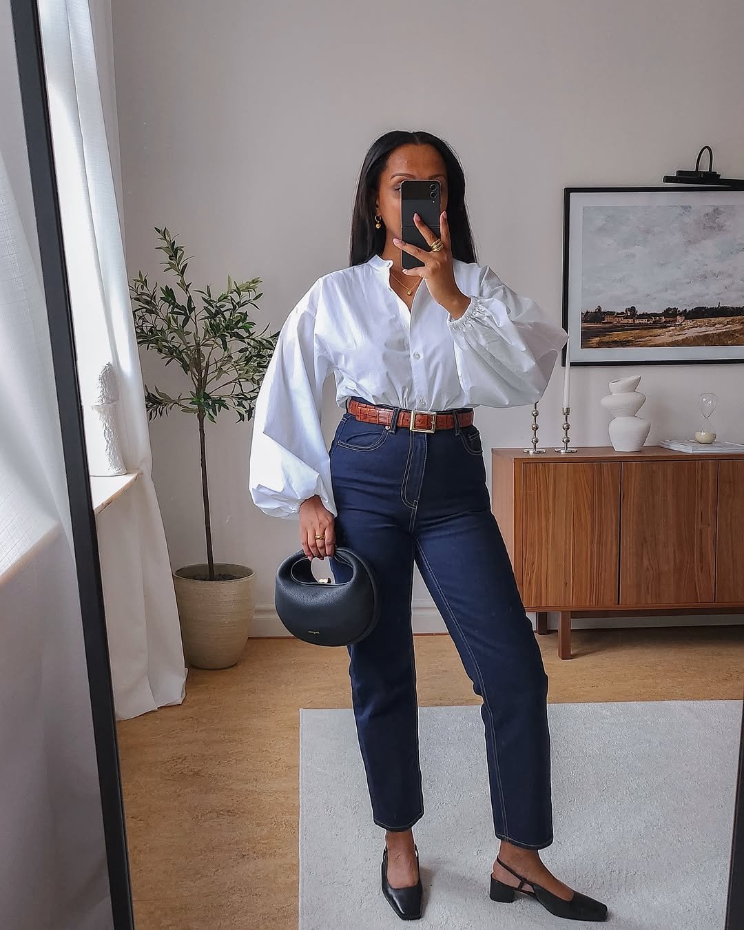 Pessoa na frente de espelho usando look com camisa branca, jeans e sapatos pretos, segurando bolsa preta redonda.