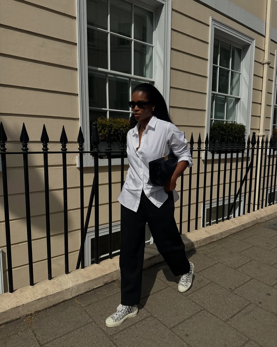 Pessoa usando look com camisa branca, calça preta e óculos escuros, caminhando em uma calçada ao lado de uma grade.