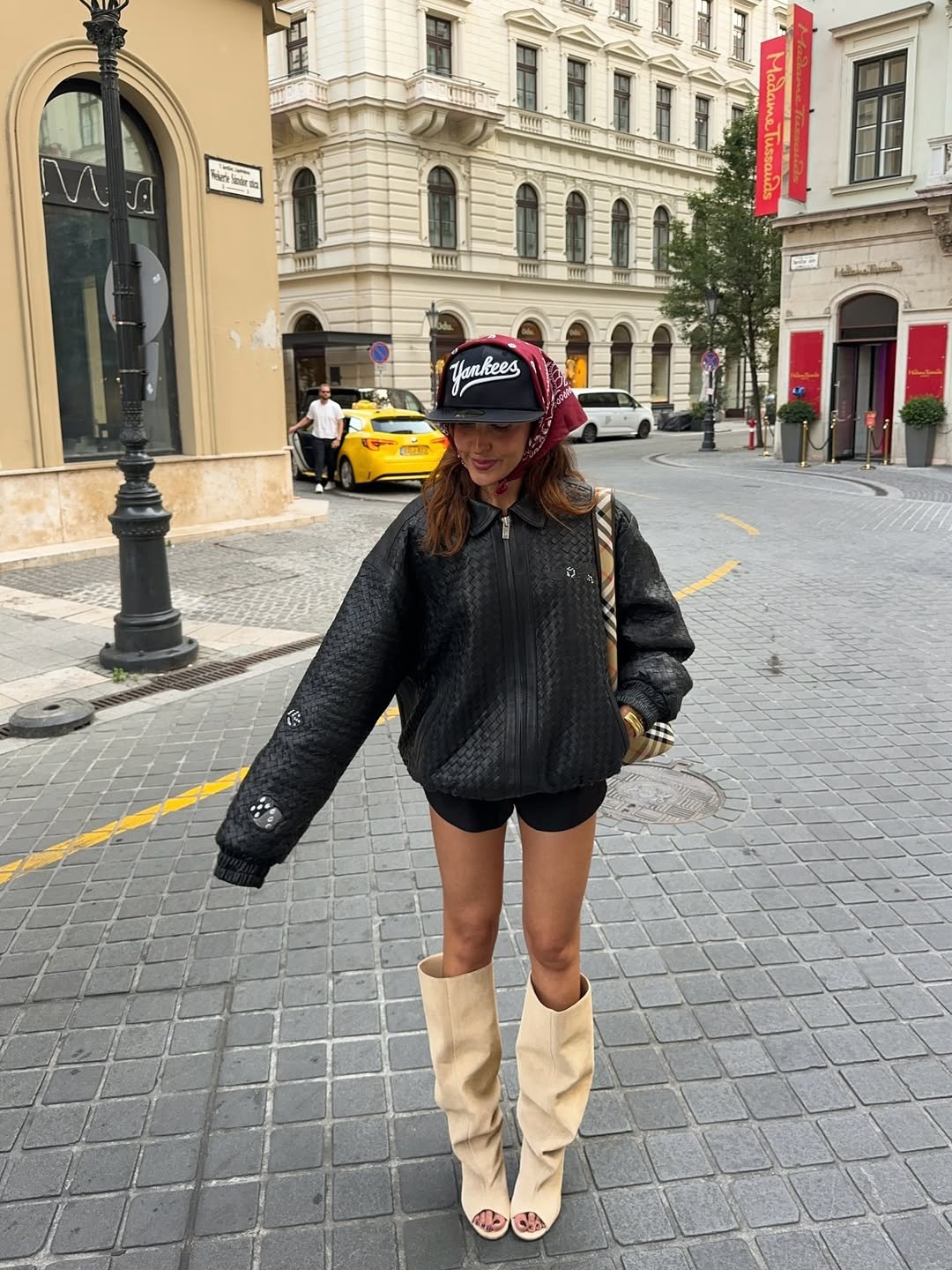 Mulher com look urbano: jaqueta preta, botas bege, lenço vermelho no boné. Edifícios clássicos ao fundo.