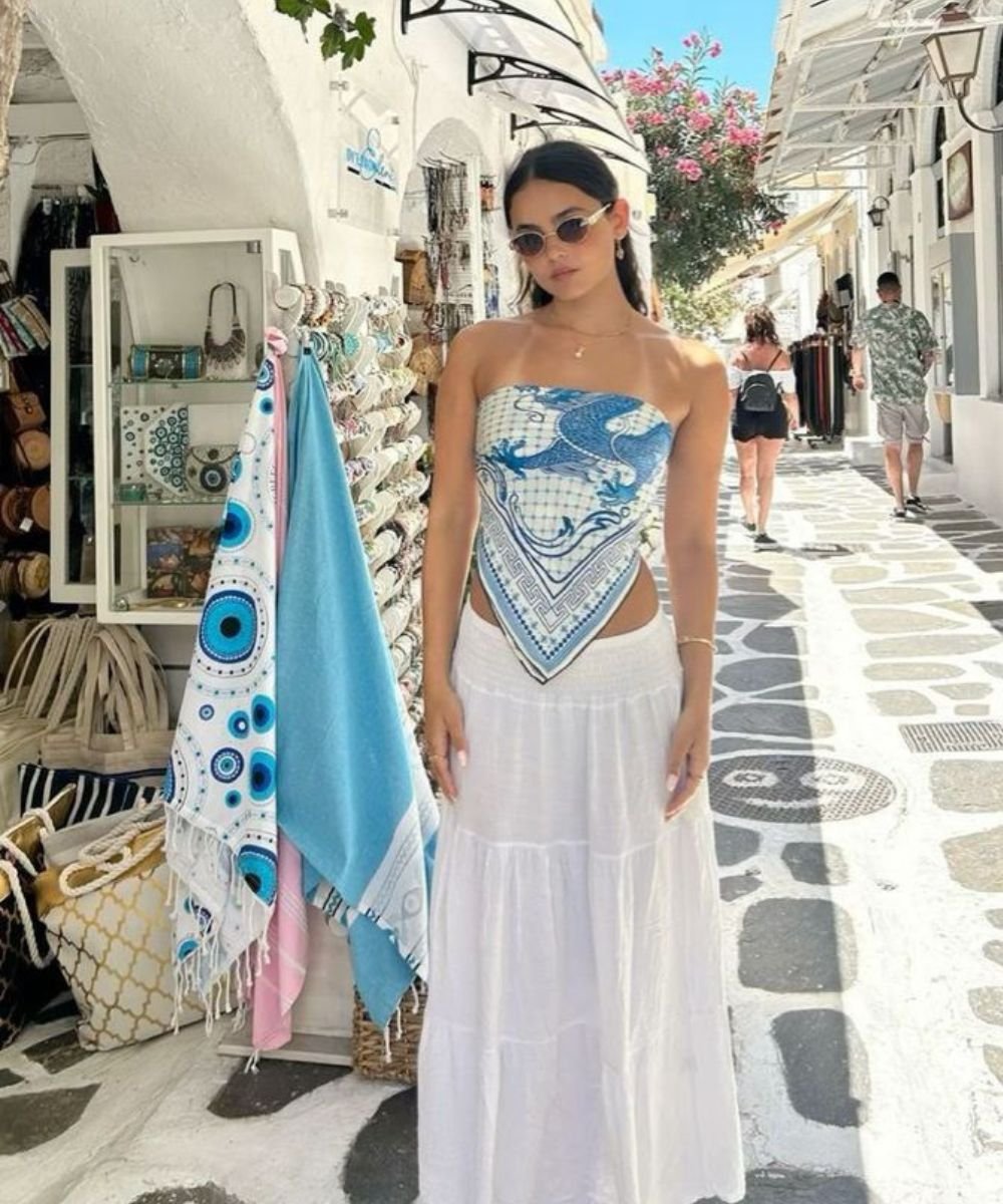 Moça em look de verão usando lenço azul como top e saia longa branca, em rua com lojas.