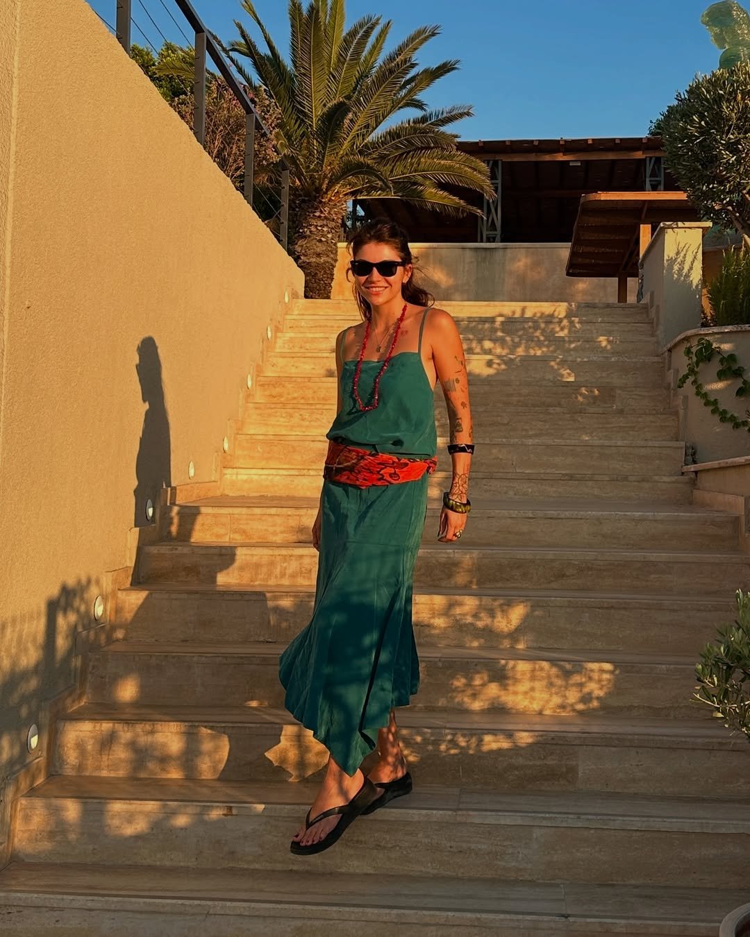 Mulher mostrando como usar lenço na cintura com vestido verde e colar vermelho ao pôr do sol.