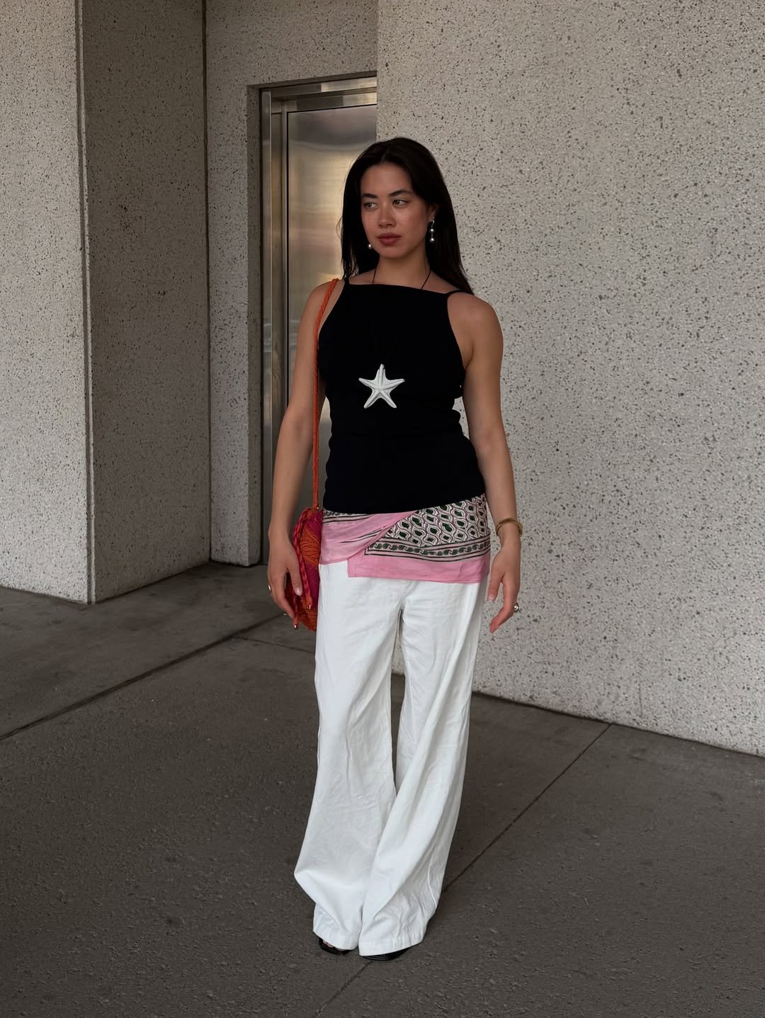 Mulher em look estiloso com lenço na cintura, blusa preta com estrela e calça branca.