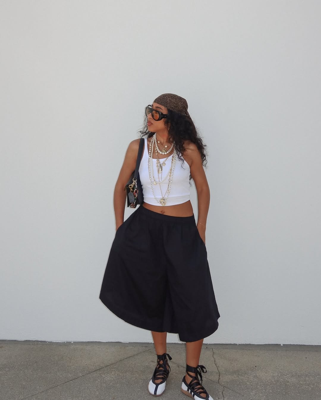 Mulher ensinando como usar lenço na cabeça. Look com top branco, saia larga preta e colares.