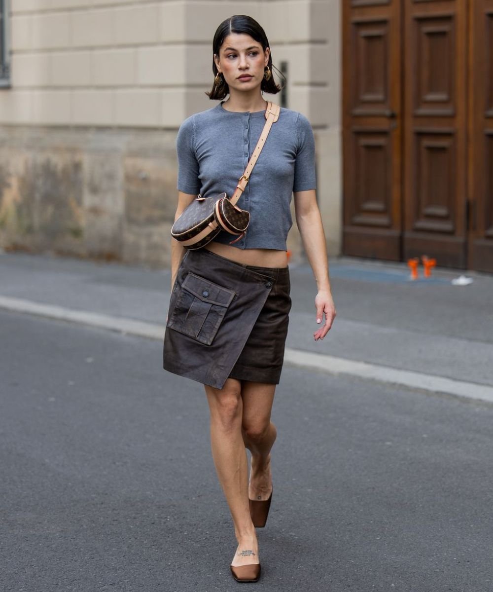 Mulher usando look com saia envelope marrom, blusa cinza e bolsa transversal marrom.