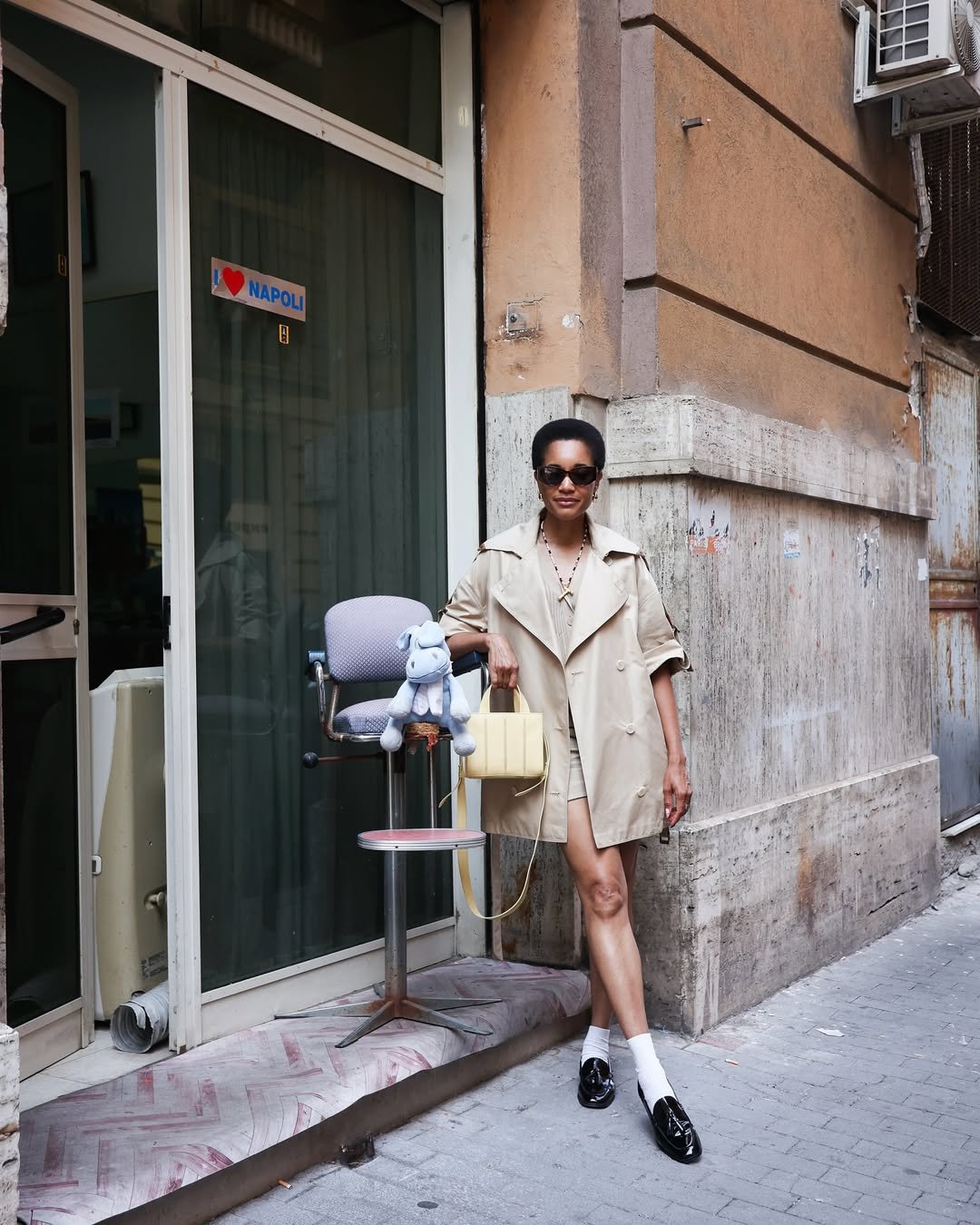 Mulher de óculos escuros usa casaco bege, apoiada em cadeira com boneco de pelúcia. Bolsa elegante na mão. Look comfy chique