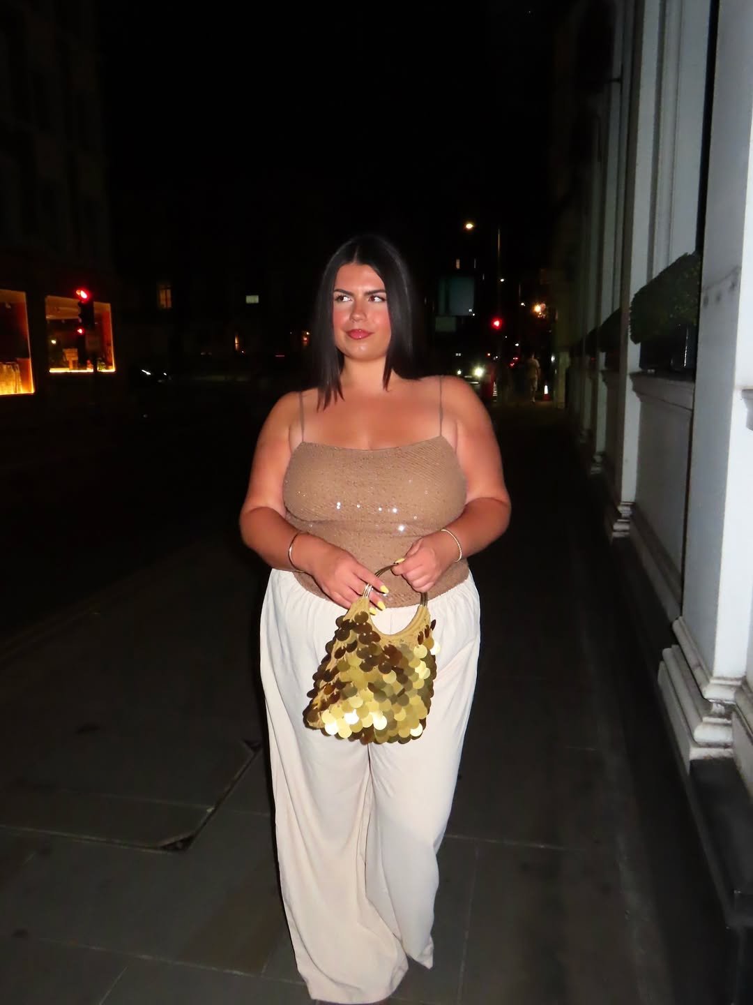 Pessoa à noite, usando blusa brilhante e calças largas, segurando bolsa dourada. Visual elegante e confortável. Look comfy para noite