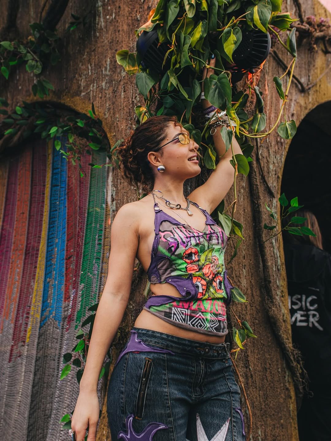 Pessoa com look de festival colorido, sorrindo e levantando o braço próximo a árvore decorada com folhas verdes.
