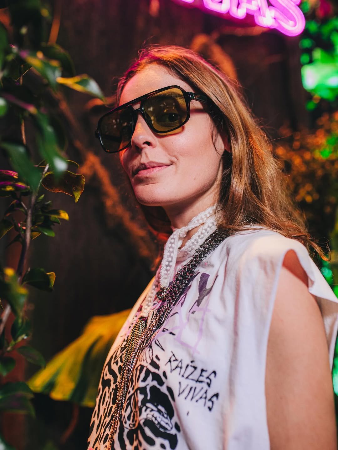 Pessoa com óculos de sol, roupa estampada e colar, em ambiente iluminado, em look de festival.