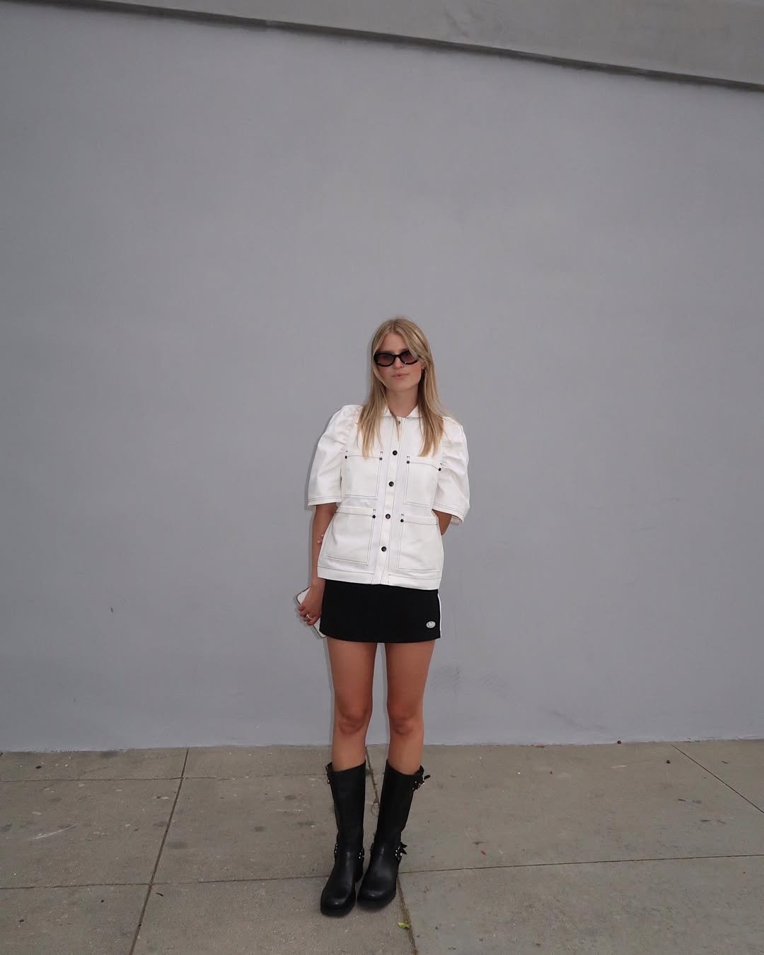 Pessoa com look de festival, usando jaqueta branca, saia preta, botas e óculos escuros em frente a uma parede cinza.