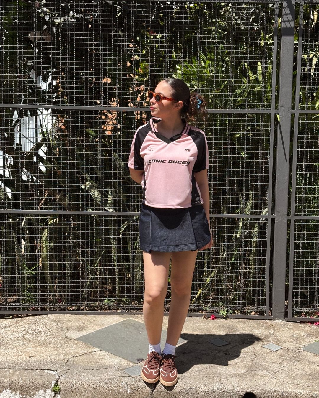 Pessoa com camiseta esportiva rosa e saia preta, óculos de sol, em frente a uma grade, aproveitando looks de primavera estilosos.