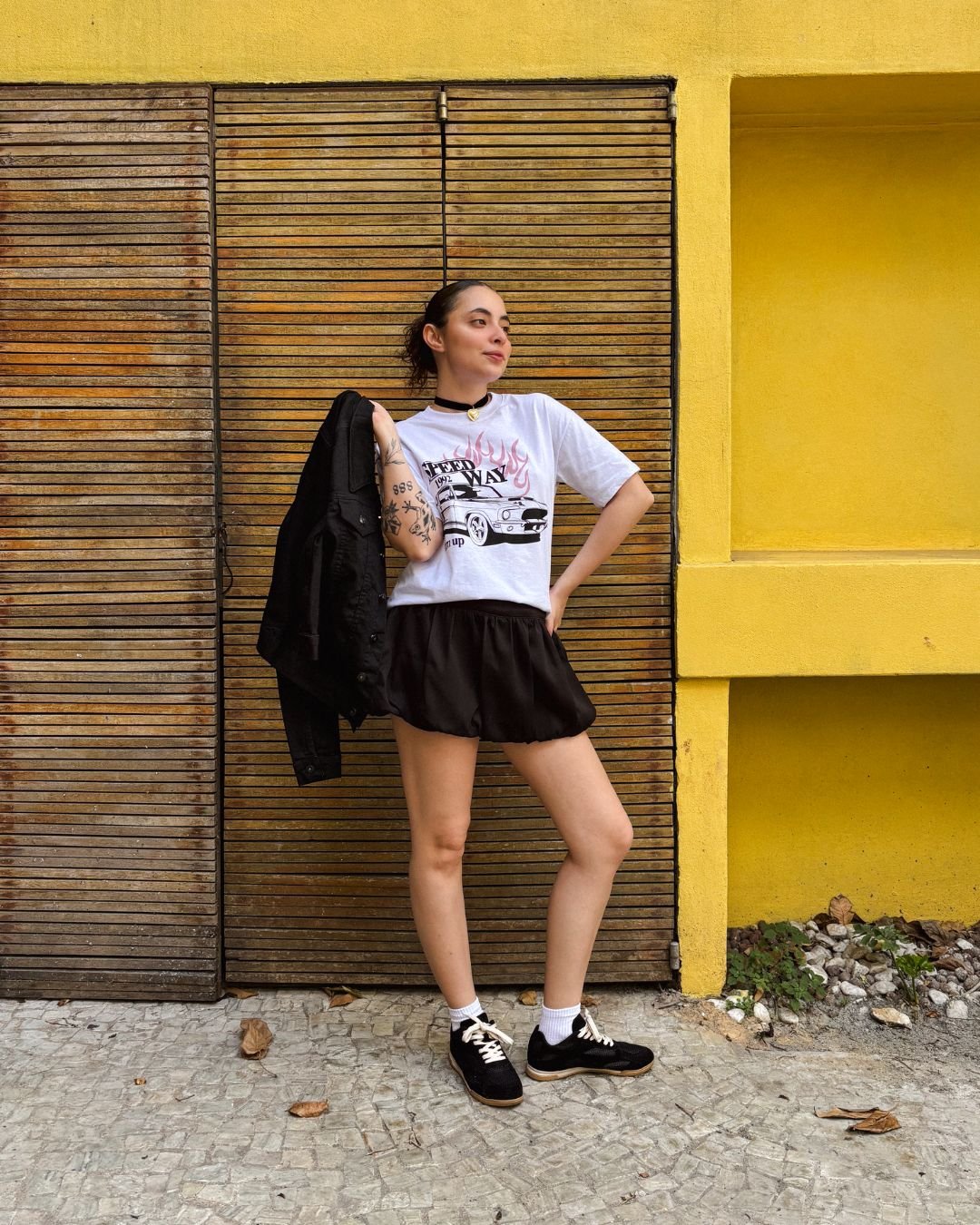 Vestida com camiseta gráfica e shorts pretos, segurando jaqueta. Combinação casual destaca looks de primavera estilosos.