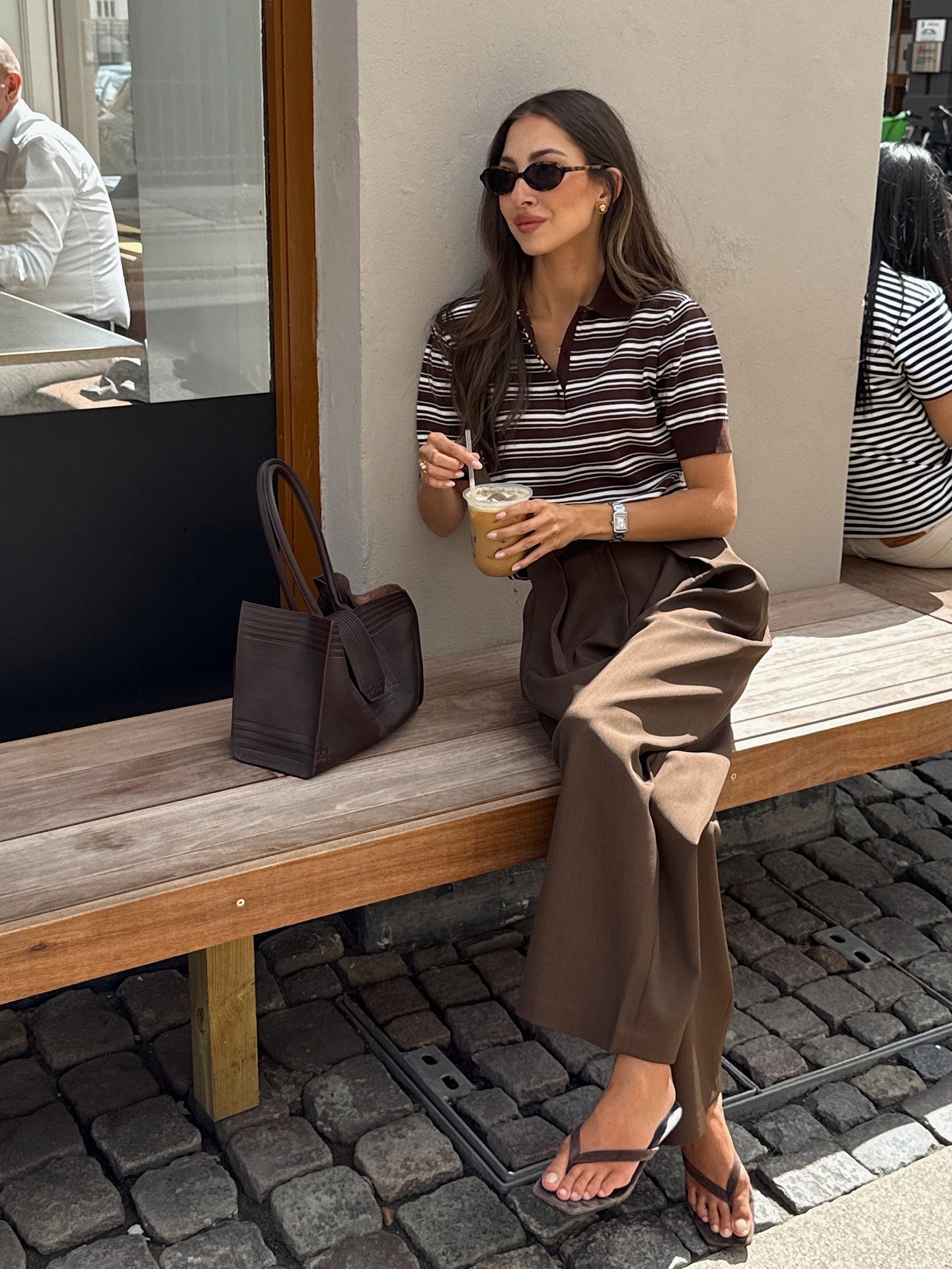 Mulher sentada em banco usando óculos escuros e roupa marrom listrada, segurando bebida. Looks bonitos e elegantes.