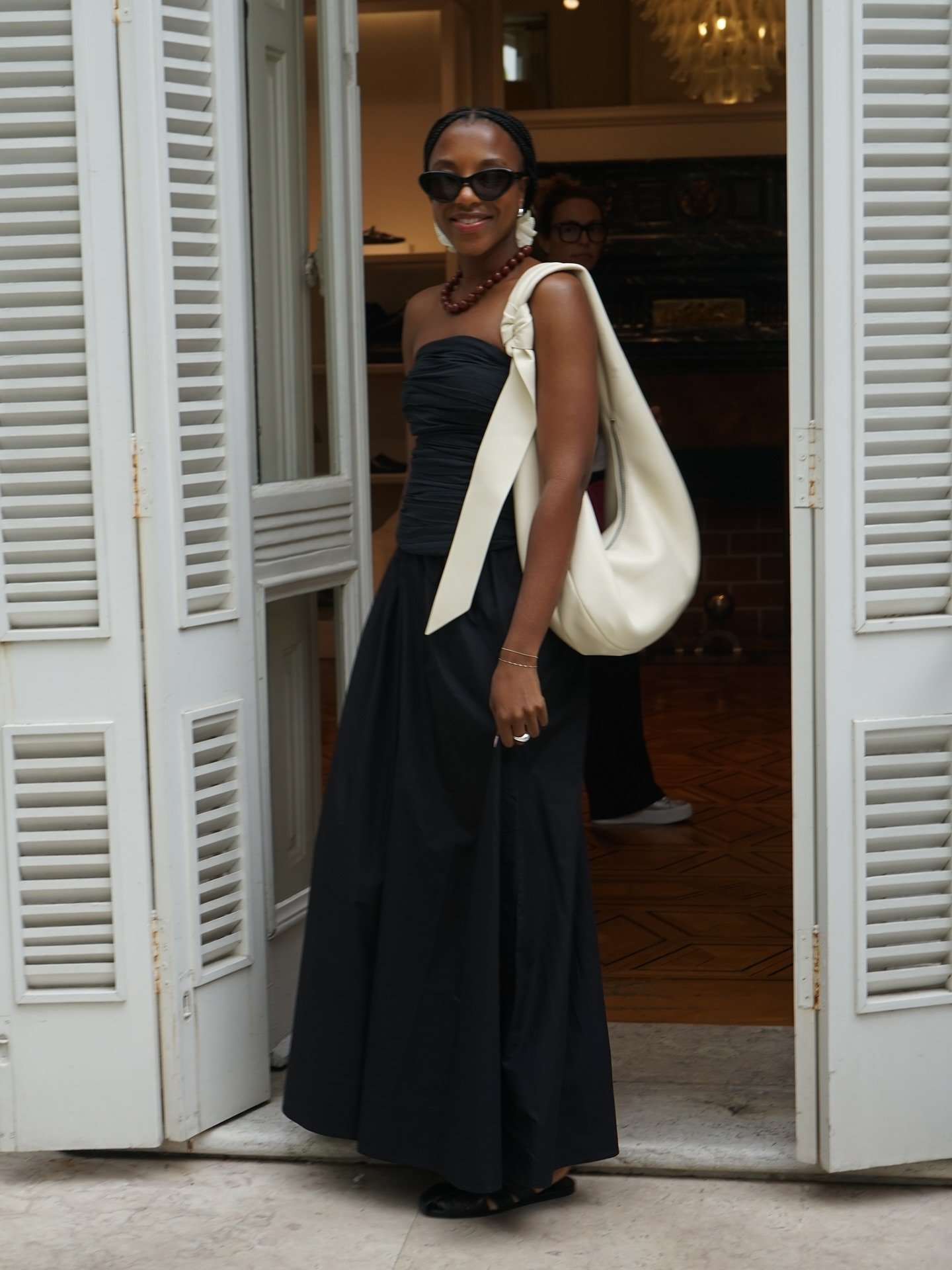 Pessoa sorrindo com vestido preto elegante e bolsa branca, exibindo looks bonitos ao sair de uma loja.