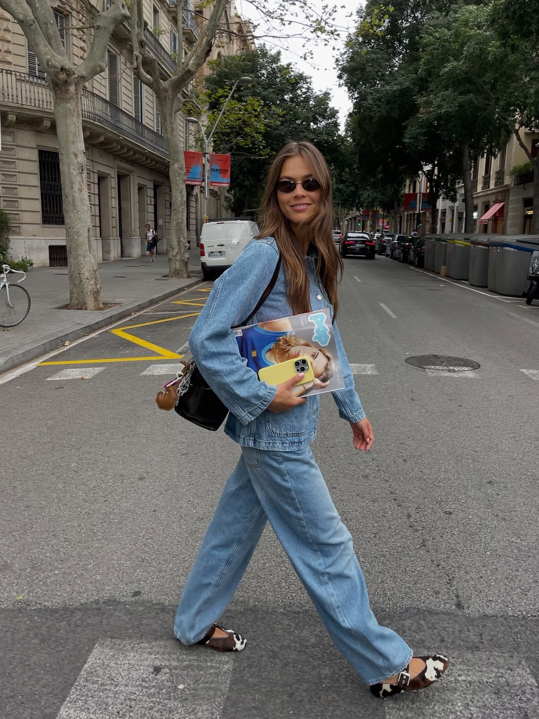 Pessoa caminhando na rua com conjunto jeans e óculos escuros. Carrega revista e celular. Looks bonitos.