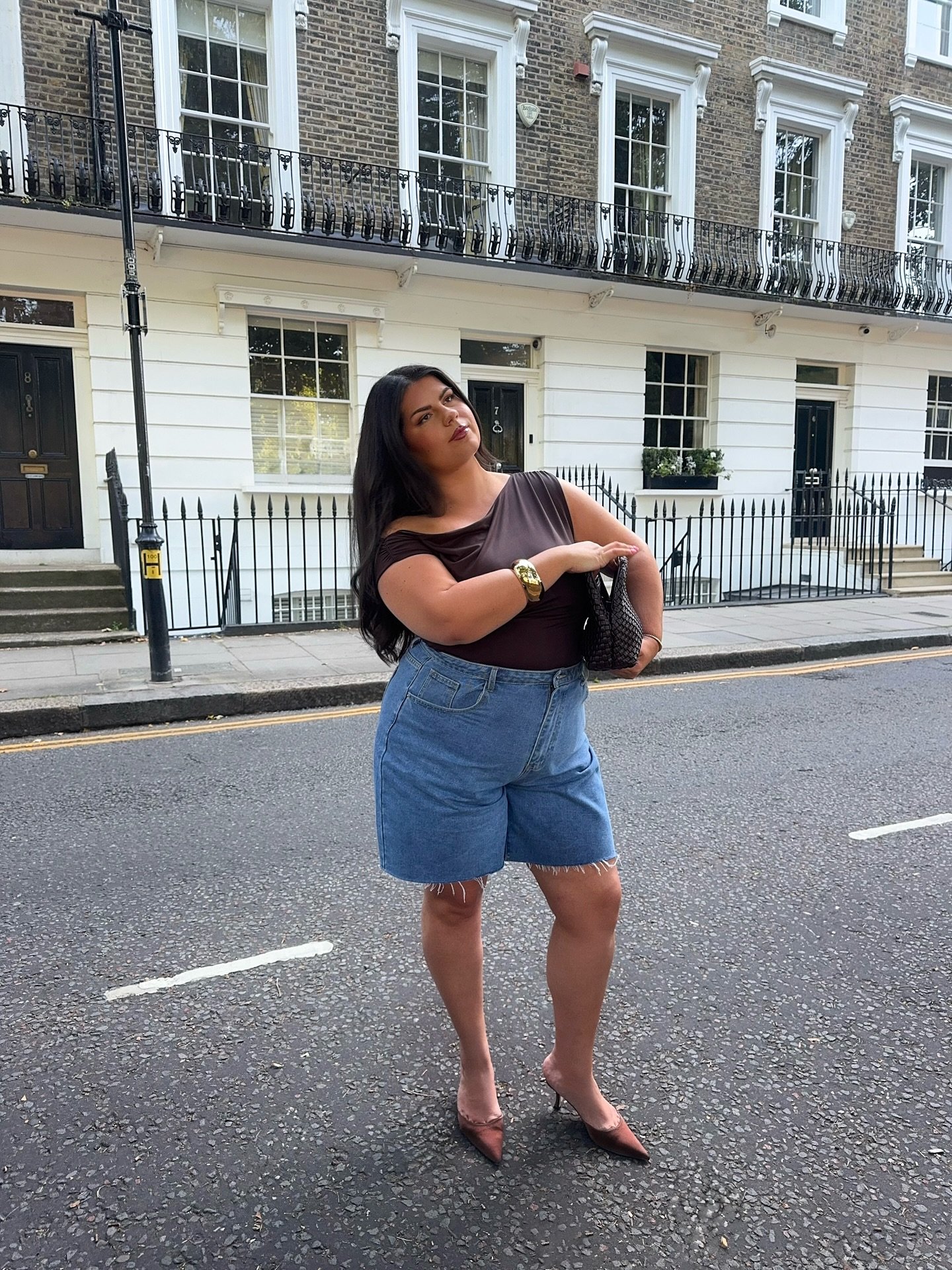 Mulher com camiseta marrom e shorts jeans, parada em rua de bairro com casas tradicionais.