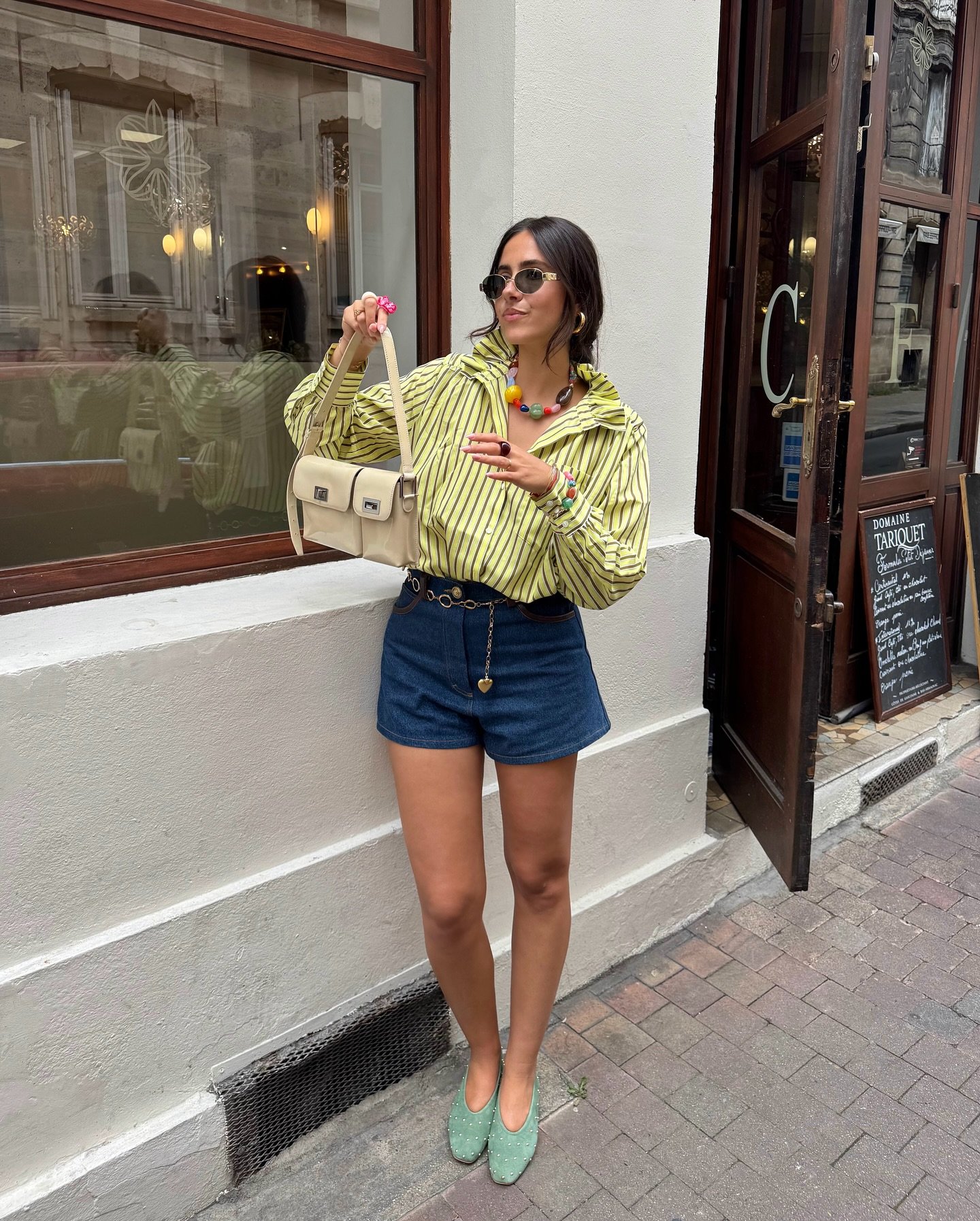 Pessoa com blusa listrada e shorts jeans em frente a um café, exibindo looks bonitos e modernos.