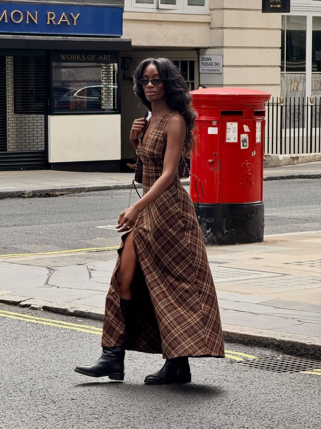 Mulher com vestido xadrez longo e botas caminha em rua urbana, próximo a uma caixa de correio vermelha.